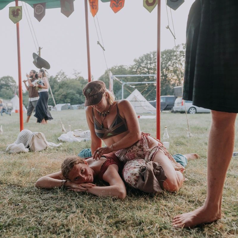 One participant caring for another lying on the grass under a tent during an outdoor gathering.