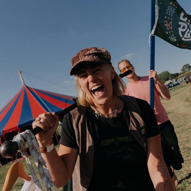 Person wearing a cap holding a staff beside a flag near a large outdoor tent.